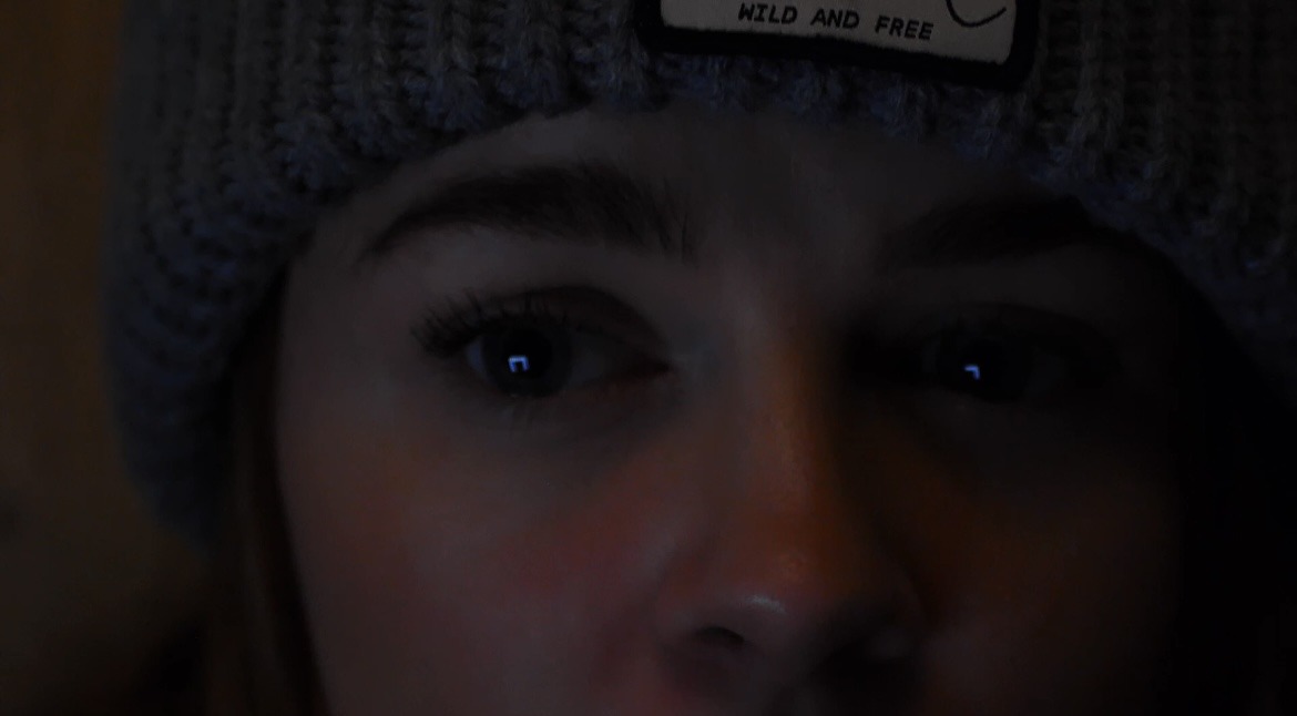 A woman in a knitted beanie lit by the blue glow of a screen
